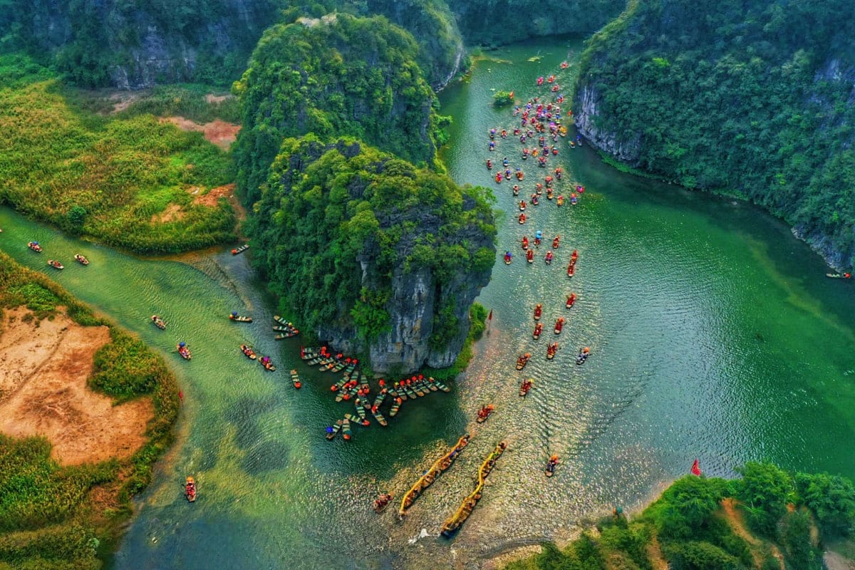 Vietnam Cultural Journey for Families Aerial view of tourists in red kayaks on emerald river surrounded by towering limestone cliffs and lush green vegetation in H