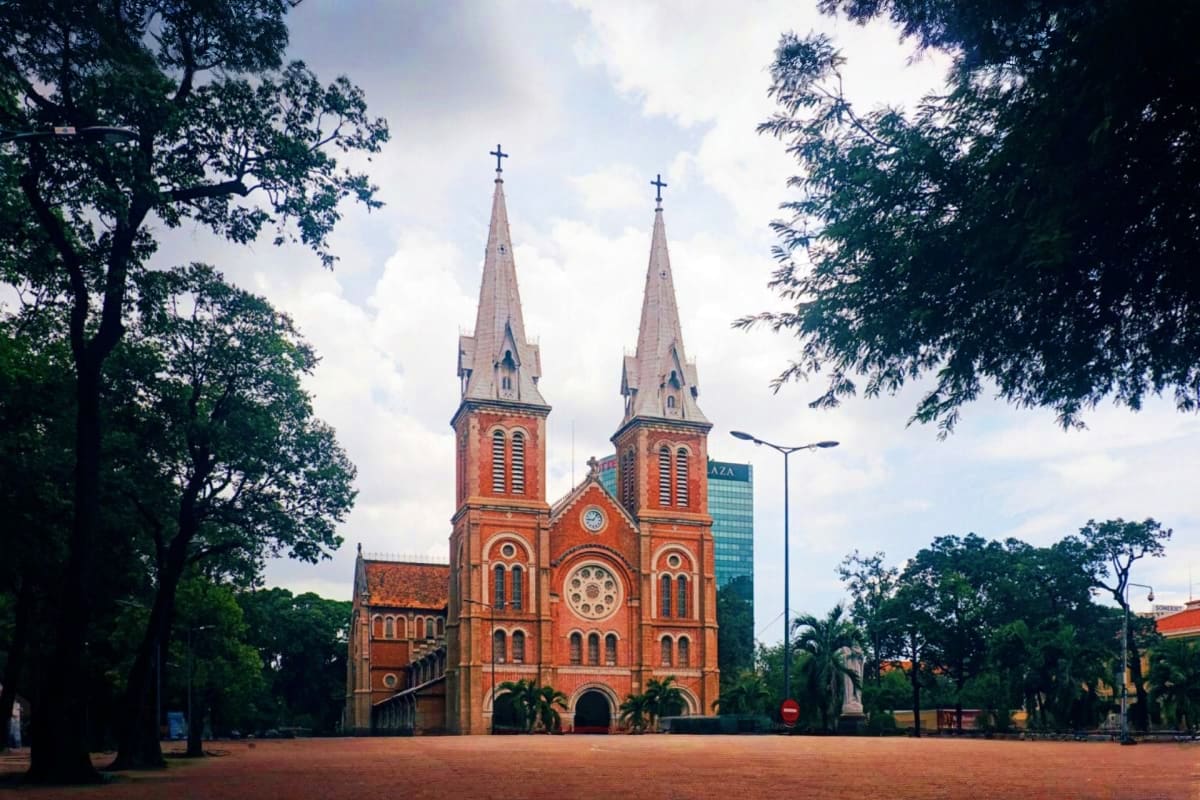 Vietnam Cultural Journey for Families Historic red brick cathedral with twin spires and cross towers surrounded by lush green trees in Ho Chi Minh City, Vietnam