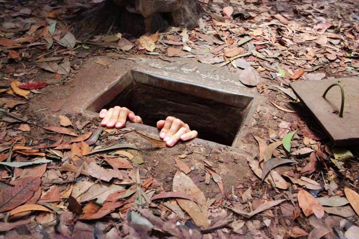 Vietnam Cultural Journey for Families Hands emerging from underground tunnel opening in soil covered with fallen leaves in Vietnamese countryside