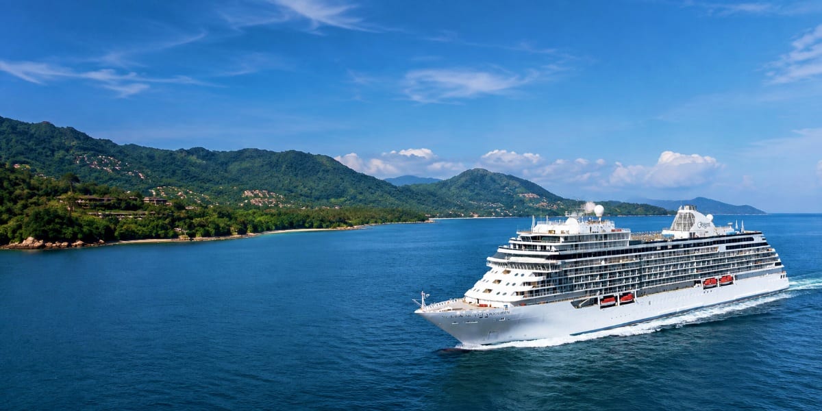 Large white cruise ship sailing in turquoise waters with lush green mountains and beach coastline in background under blue sk