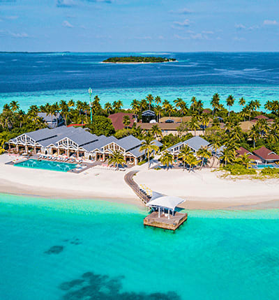 Aerial view of a tropical resort with white buildings, swimming pool, and wooden dock extending into turquoise lagoon with di