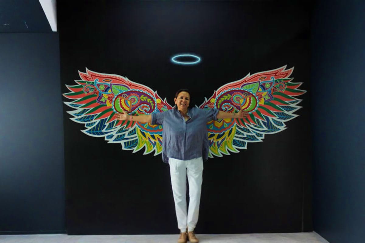 Woman posing with arms outstretched in front of colorful 3D angel wings mural with glowing halo at interactive museum install