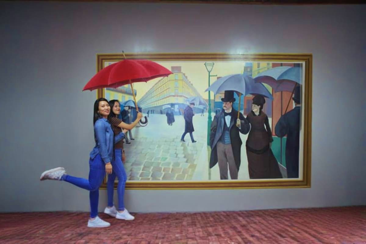 Two visitors holding a red umbrella posing in front of a 3D painted mural of a rainy European street scene at the Selfie Muse
