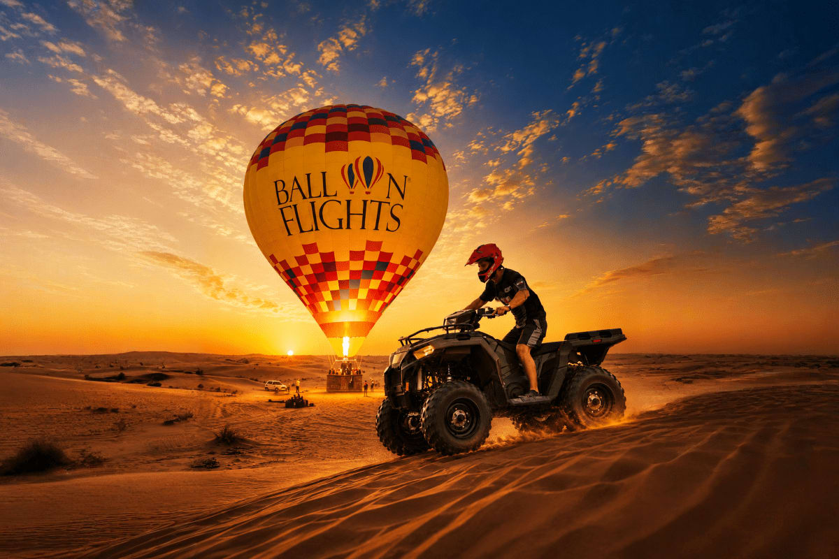 Red and yellow checkered hot air balloon labeled "Balloon Flights" at sunrise with rider on ATV in desert landscape