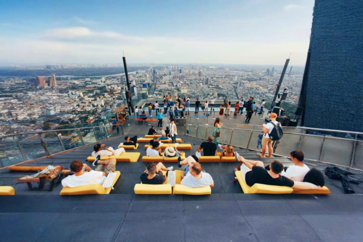 Visitors lounging on yellow cushioned seating on the Mahanakhon SkyWalk observation deck with panoramic Bangkok cityscape vie