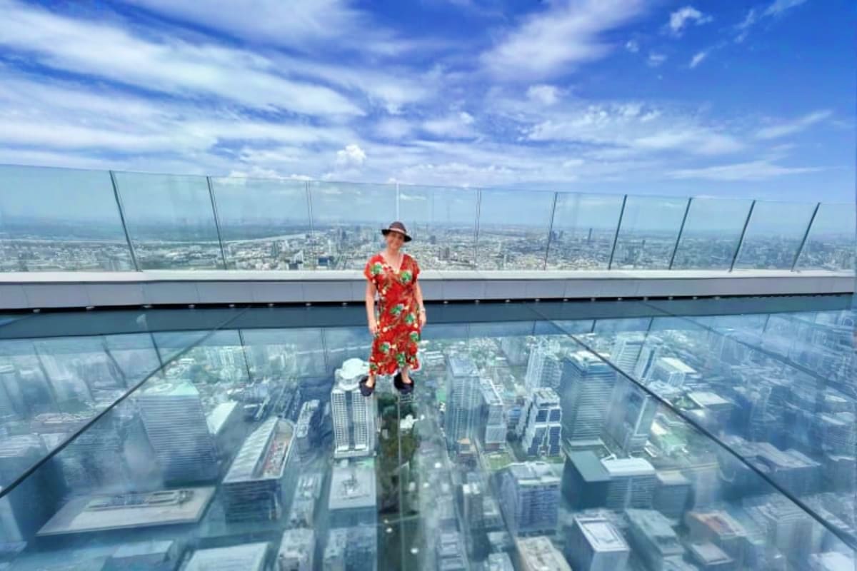 Woman standing on glass floor at Mahanakhon Skywalk observation deck with Bangkok cityscape view below and blue sky above