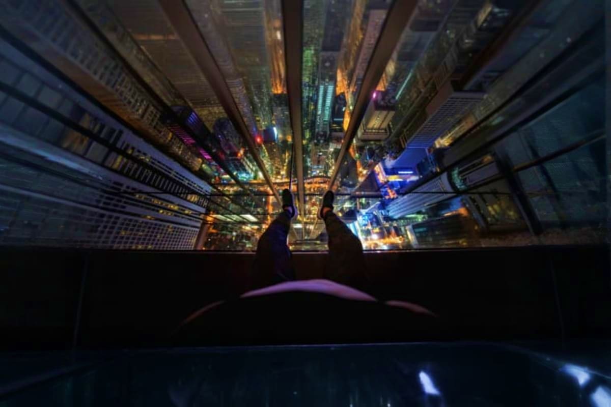 First-person view of feet on glass floor of Mahanakhon Skywalk with illuminated Bangkok cityscape and skyscrapers visible bel