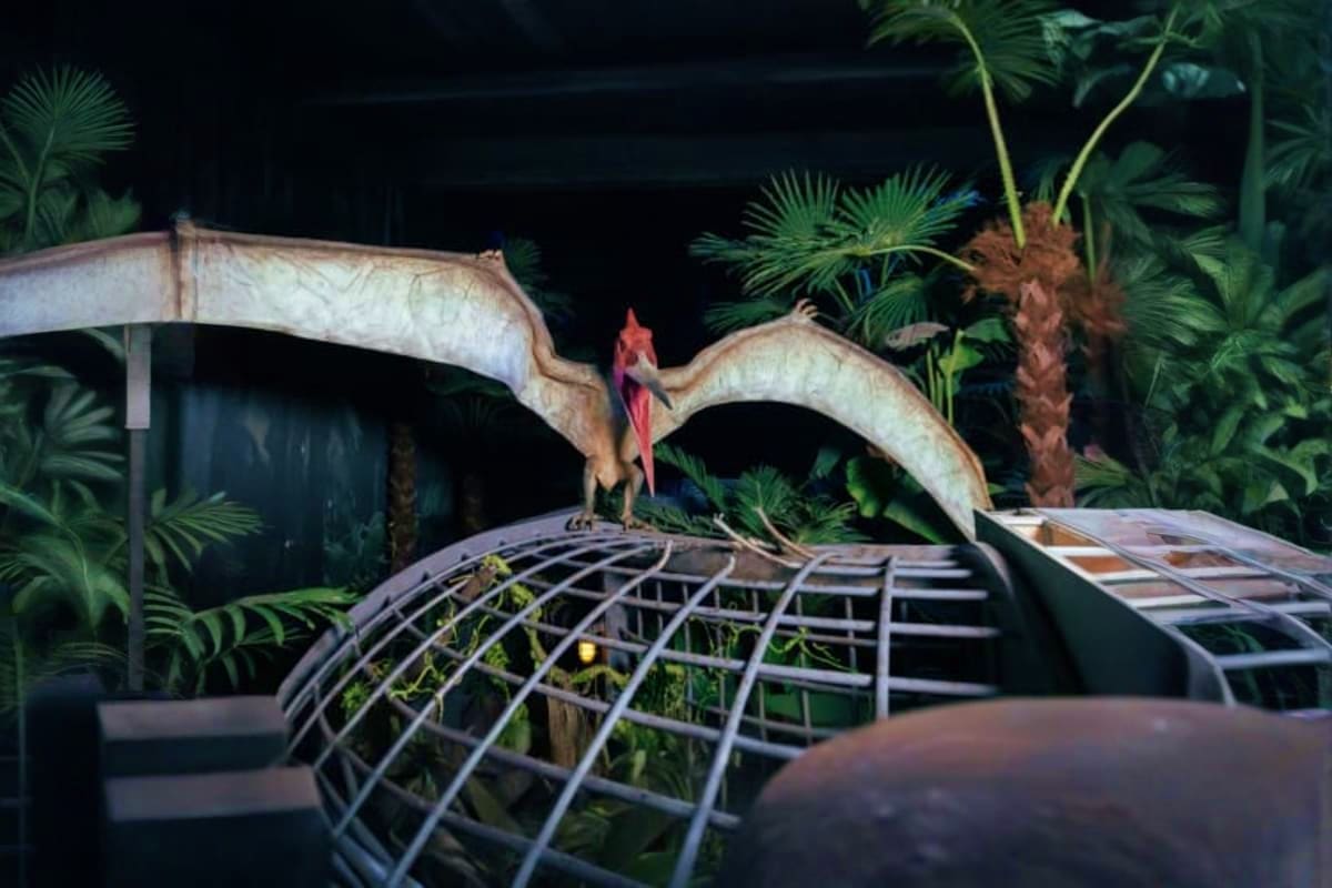 Dinosaur walking across curved bridge structure in tropical jungle setting with palm trees and metal railings at Jurassic Wor