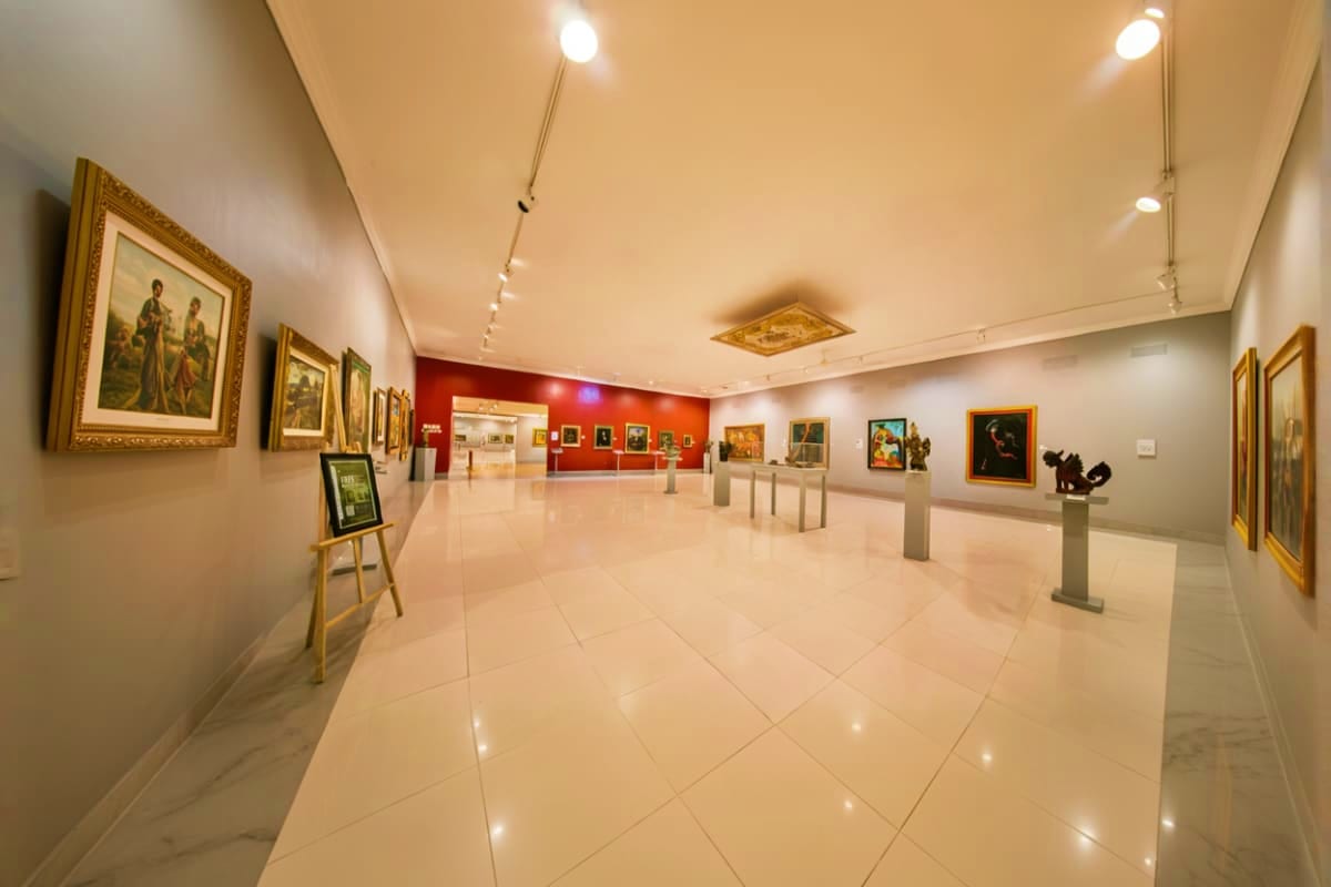 Museum Pasifika Bali Modern museum gallery interior with polished tile floors, framed paintings in gold frames on white and red accent walls, trac
