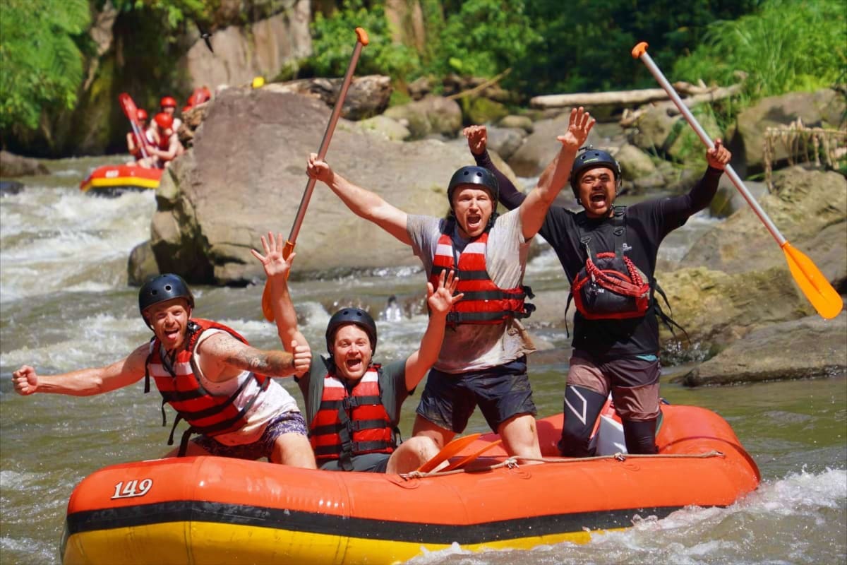 Group of five people white water rafting on orange and yellow raft in rocky river with paddles raised in celebration