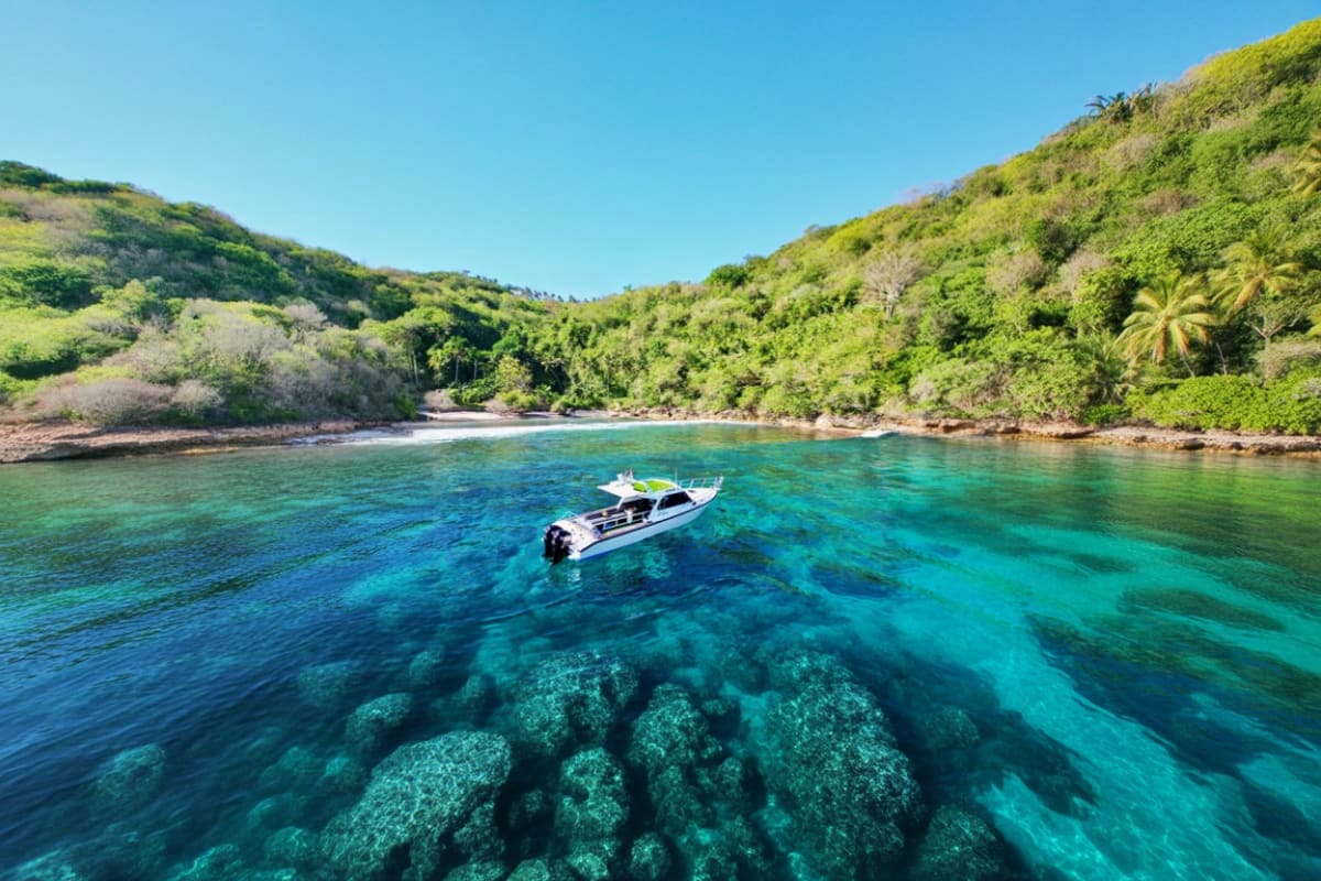 Nusa Penida Manta Bay Snorkeling Experience Motorboat anchored in crystal-clear turquoise water with visible coral reef below, tropical hillside with lush green vegetati