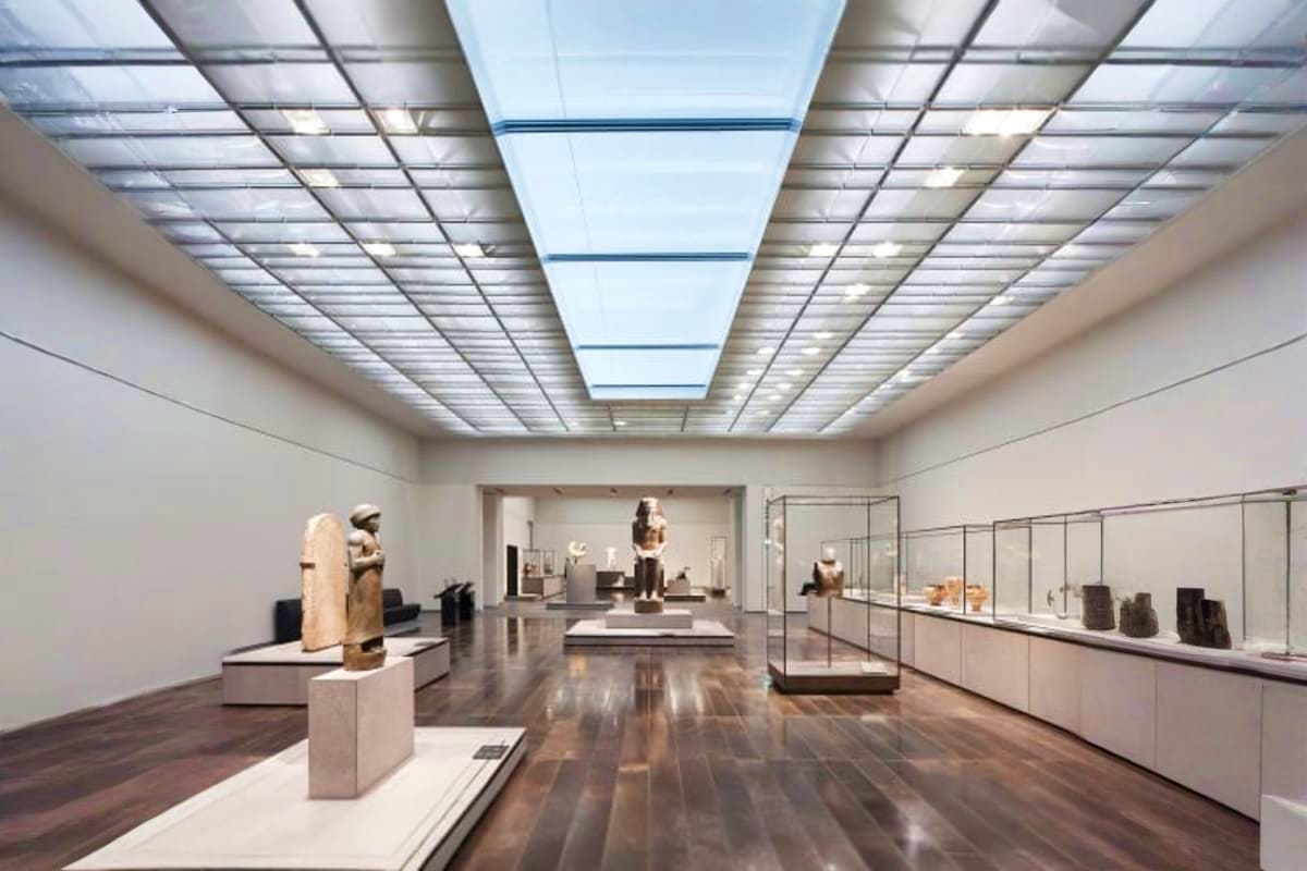 Modern museum gallery interior with ancient Egyptian sculptures displayed on pedestals, skylights, and glass exhibition cases