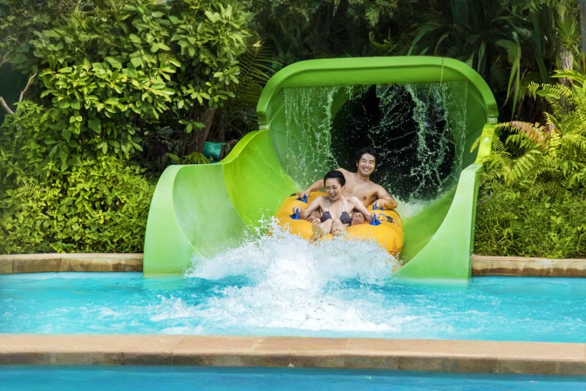 Two people riding yellow tube down bright green water slide splashing into turquoise pool surrounded by tropical plants