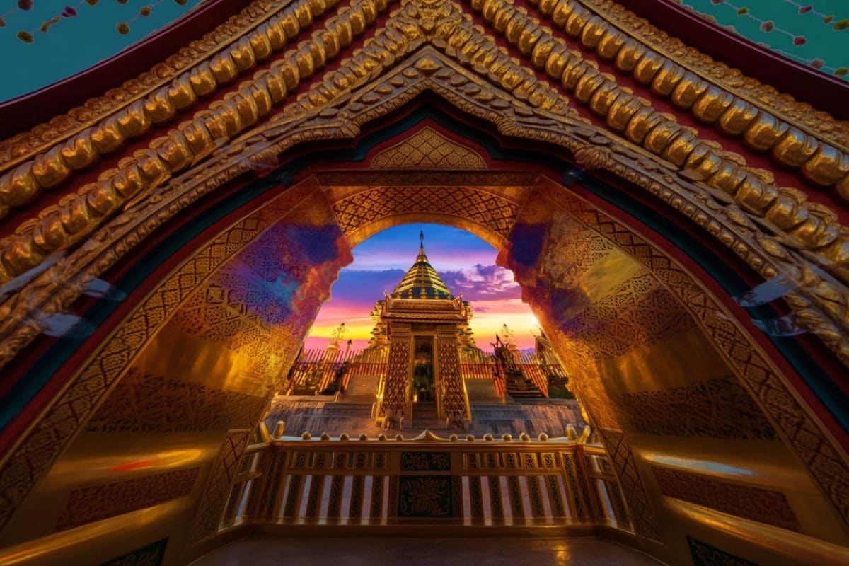 View through ornate golden archway of Doi Suthep Temple with colorful sunset sky and illuminated golden chedi in background,
