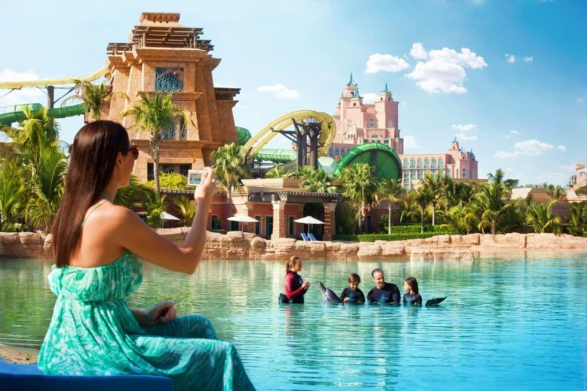 Dolphin Bay Atlantis Woman in turquoise dress photographing families swimming with dolphins in lagoon at Atlantis The Palm resort with castle and