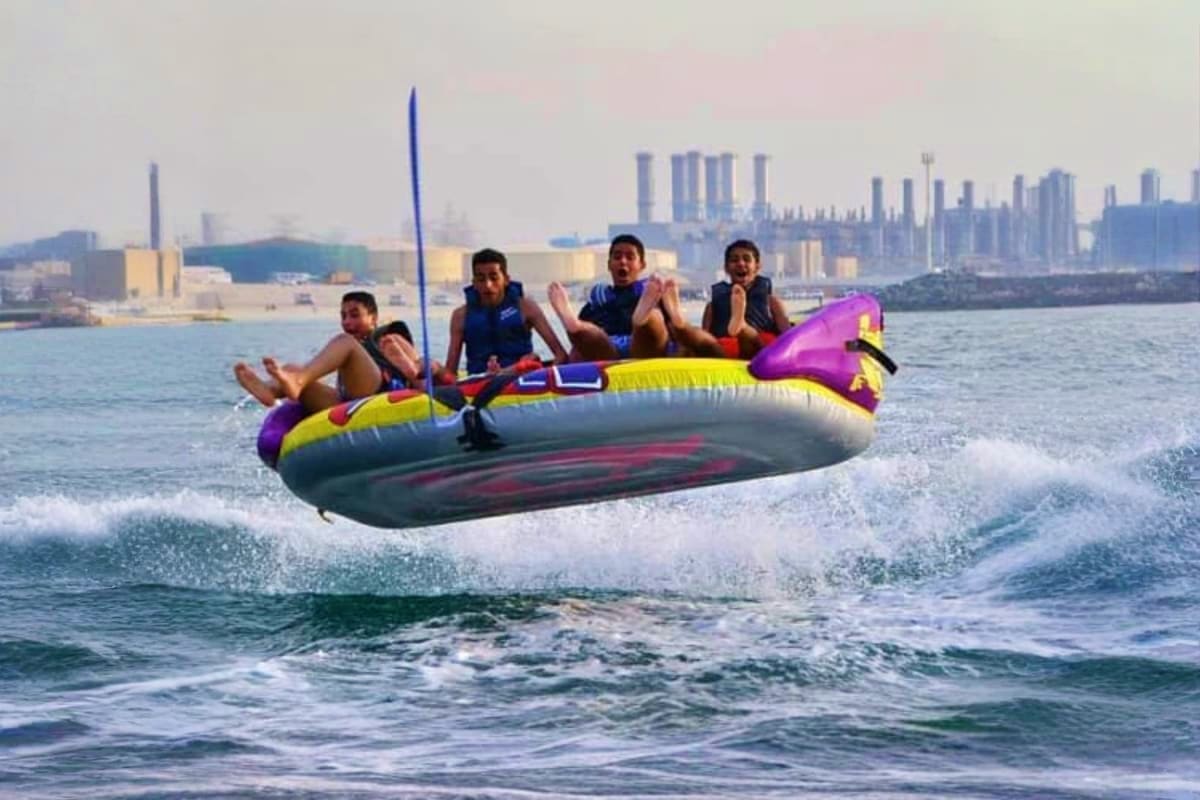 Group of people riding an inflatable donut tube in ocean waves with Dubai skyline and industrial plants visible in background