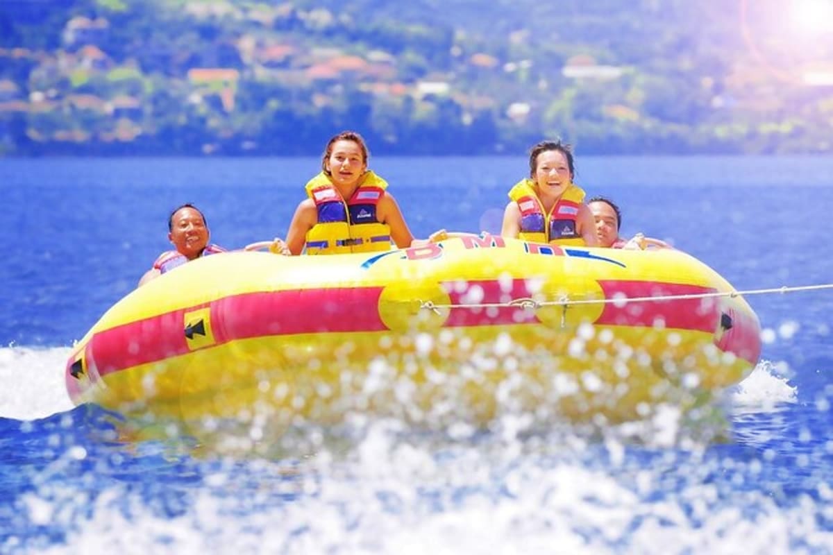 Four people wearing yellow and pink life jackets riding a yellow and red inflatable donut tube on blue ocean water with coast