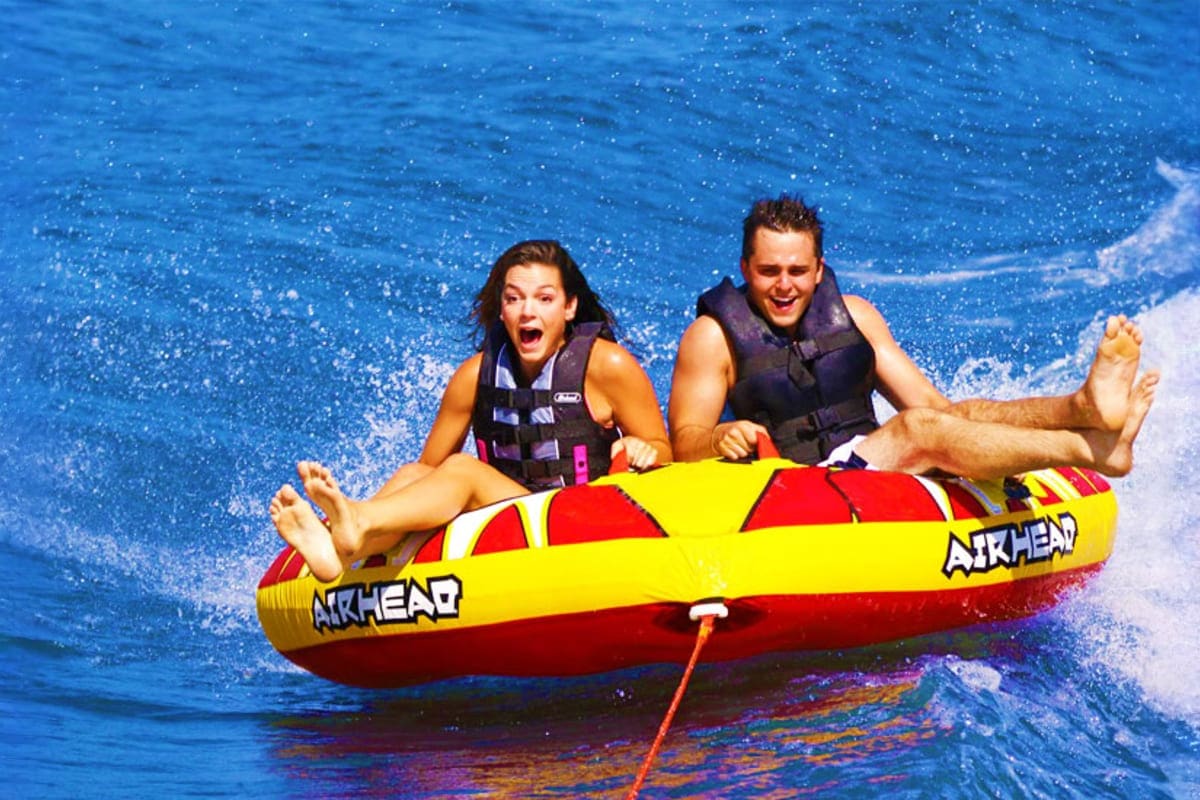 Two people wearing life jackets riding on a yellow and red inflatable donut tube being towed behind a boat in blue ocean wate