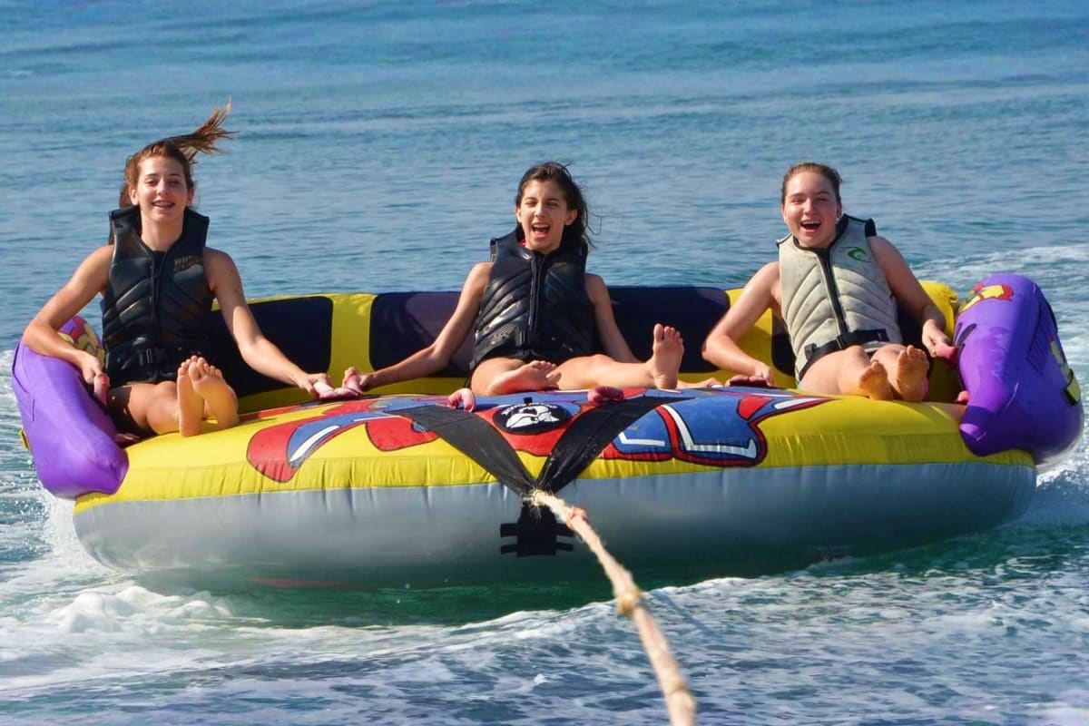 Three smiling girls sitting on a yellow and white inflatable donut tube being towed behind a boat in the ocean waves