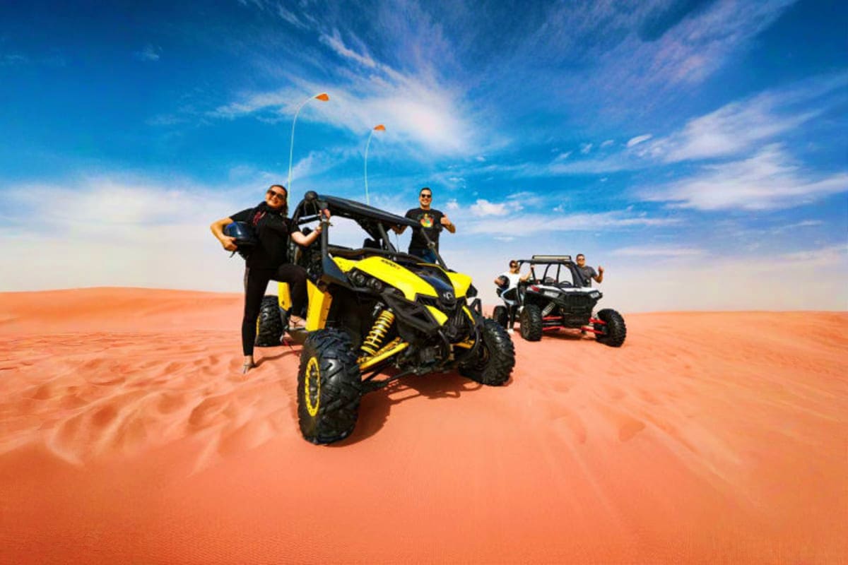 Group of tourists posing with yellow and black dune buggies on orange sand dunes under blue sky in Dubai desert