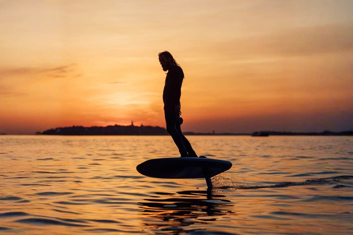 Silhouette of person standing on electric foil board in calm water at sunset with Dubai coastline in background