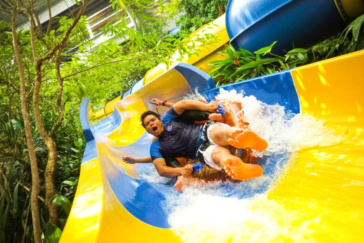 Escape Theme Park Penang Man sliding down blue and yellow water slide with splash at Escape Theme Park Penang surrounded by tropical vegetation