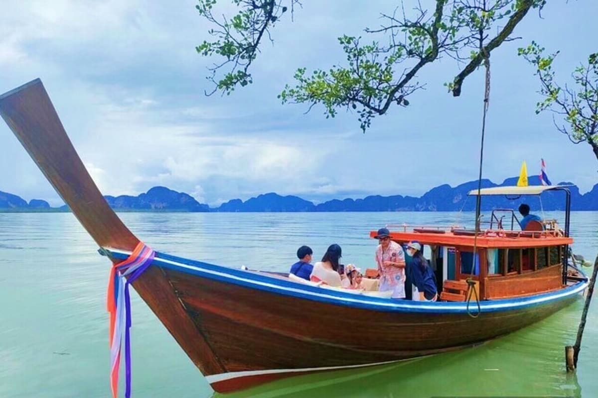Traditional wooden long tail boat with tourists moored near mangrove tree, limestone cliffs in background at James Bond Islan