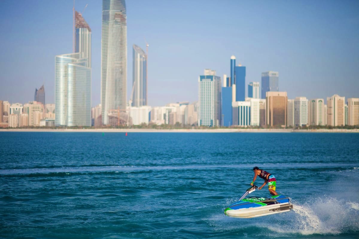 Jet ski Experience in Abu Dhabi Person riding green and blue jet ski in waves with Abu Dhabi skyline and high-rise buildings in background