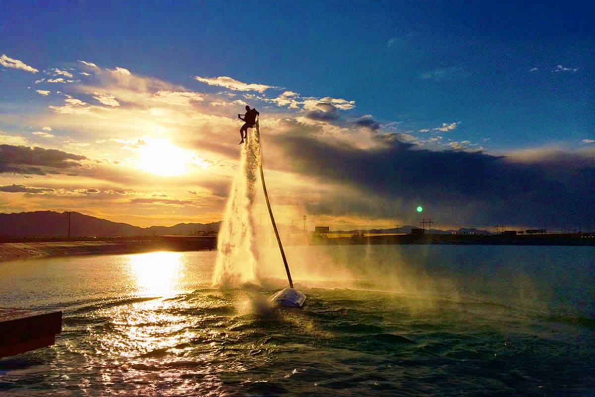 Person flying on jetpack over water at sunset with mountains and industrial structures in background