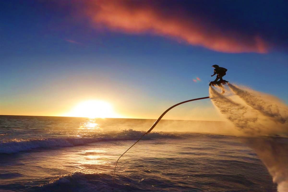 Person flying on jetpack over water at sunset with mountains and industrial structures in background (2)