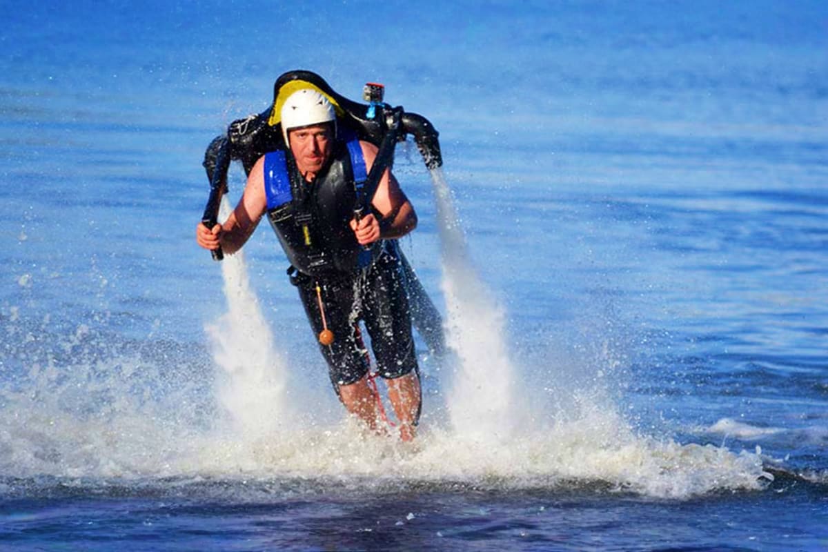 Person flying on jetpack over water at sunset with mountains and industrial structures in background (3)