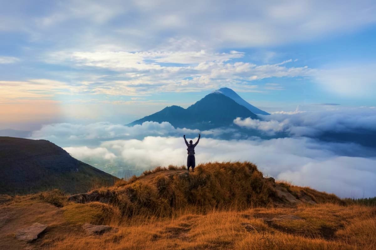 Hiker with arms raised celebrating on mountain ridge overlooking Mount Batur volcano above clouds at sunrise