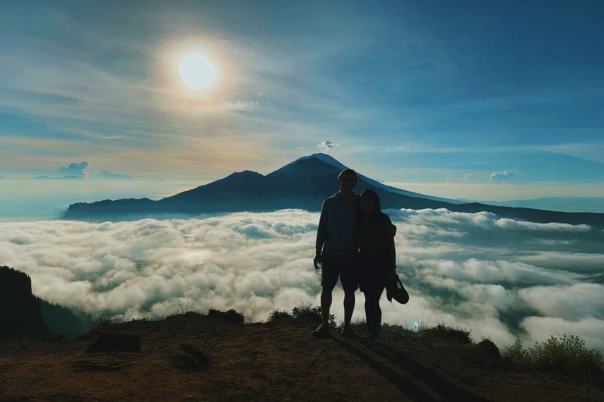 Hiker standing on mountain ridge overlooking sea of clouds with Mount Batur volcano peak at sunrise