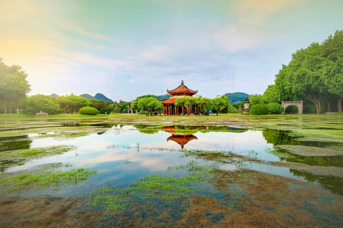 Traditional Vietnamese pagoda with orange tiled roof reflected in still water surrounded by lush green trees and mountains in