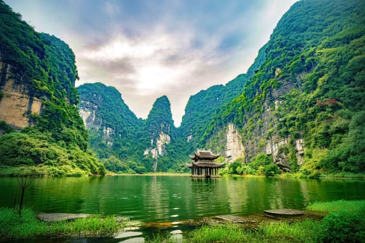 Traditional Vietnamese pagoda on emerald river surrounded by towering limestone cliffs and lush green vegetation in Ninh Binh