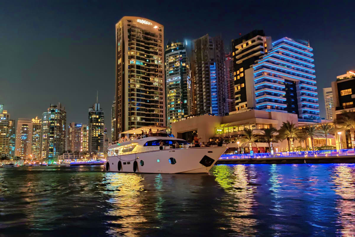 Luxury white yacht anchored at night with illuminated Dubai Marina skyscrapers and blue-lit waterfront buildings in background