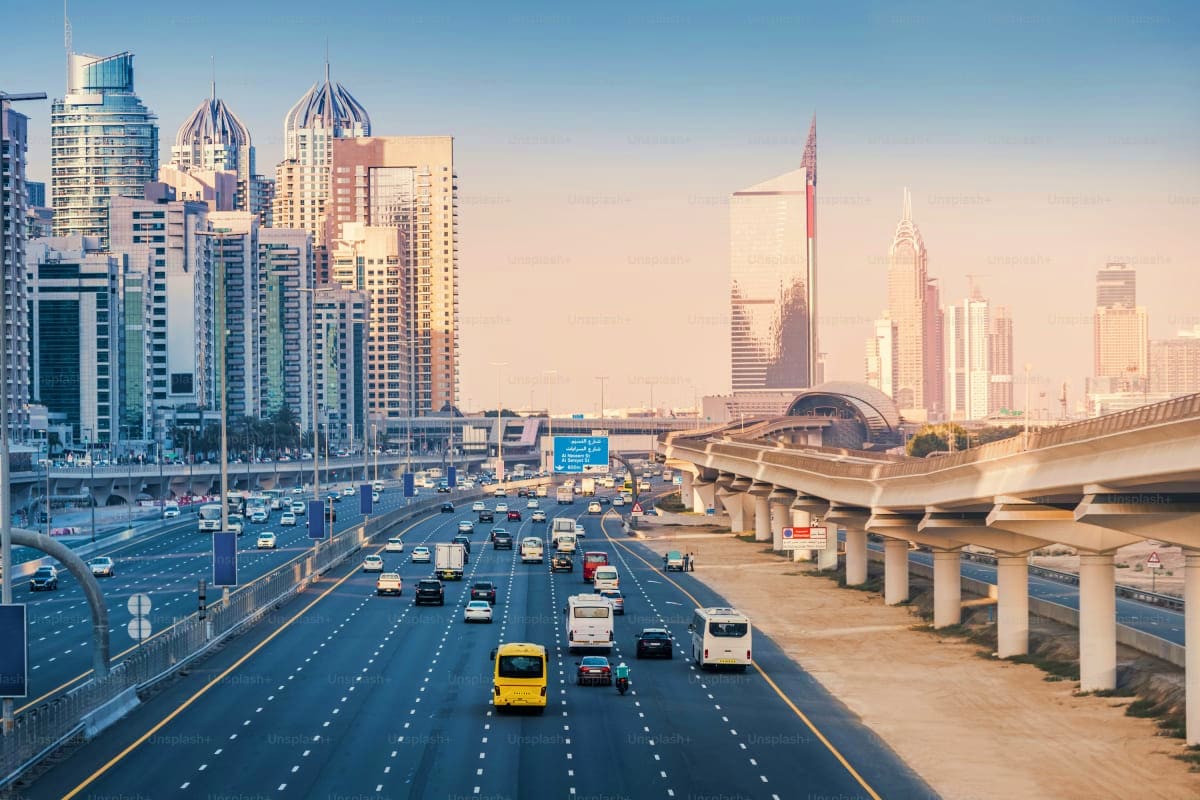 Busy Dubai highway with elevated roadway, city skyline with distinctive architecture, and vehicles including yellow taxi in b