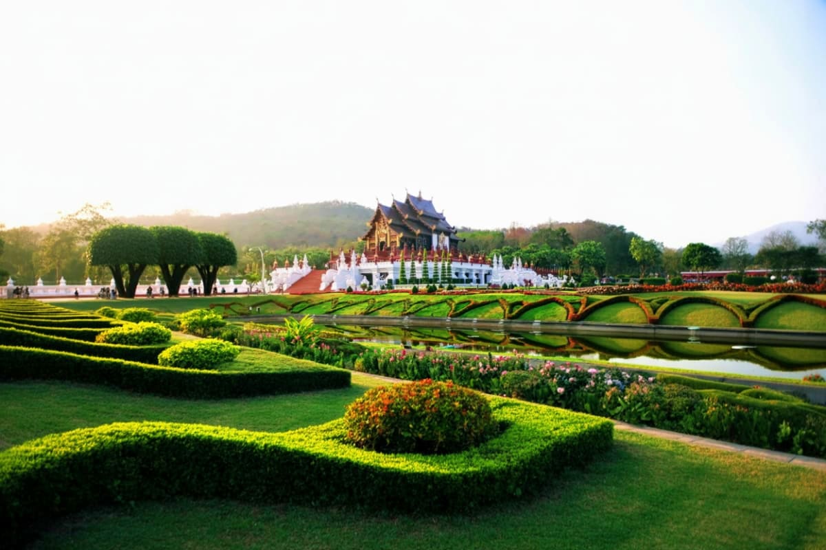 Ornate Thai temple with golden spires and white pavilion overlooking manicured gardens, topiary hedges, and wooden bridge spa
