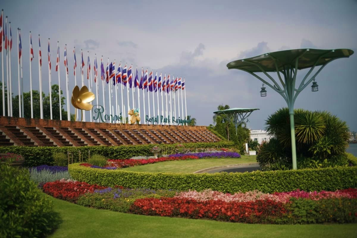 Royal Park Rajapruek entrance with international flags, manicured flower gardens in red and purple, and modern pavilion struc