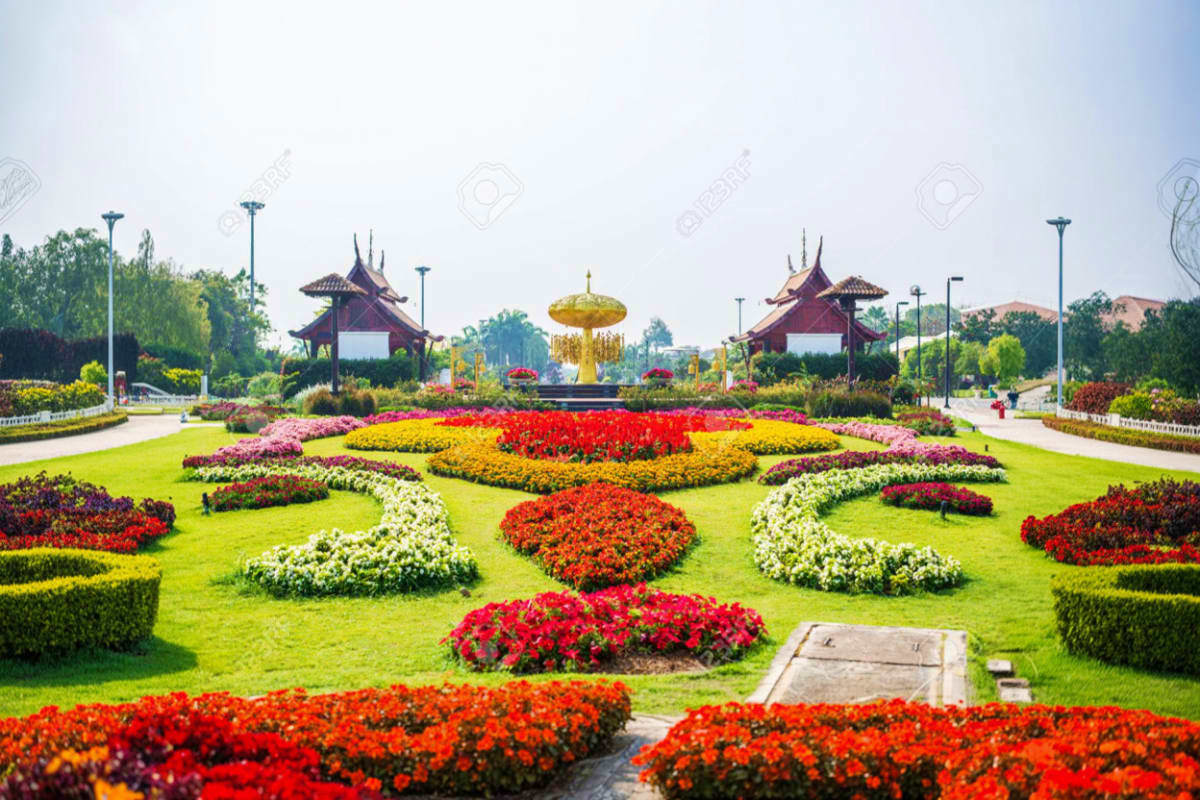 Colorful flower beds with red, yellow, and white blooms in a manicured garden with traditional Thai pavilions and golden foun