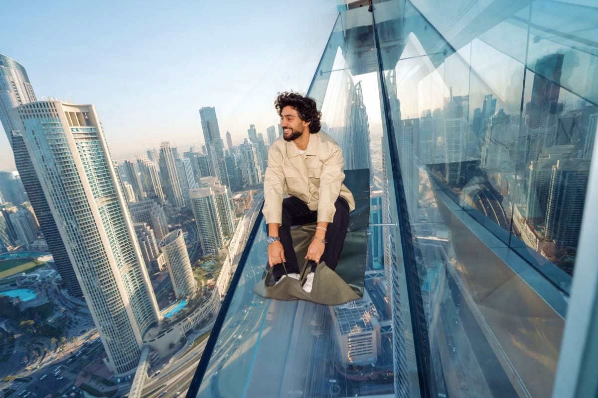 Person with arms raised sitting in glass slide overlooking Dubai cityscape and skyscrapers from high-rise building (2)