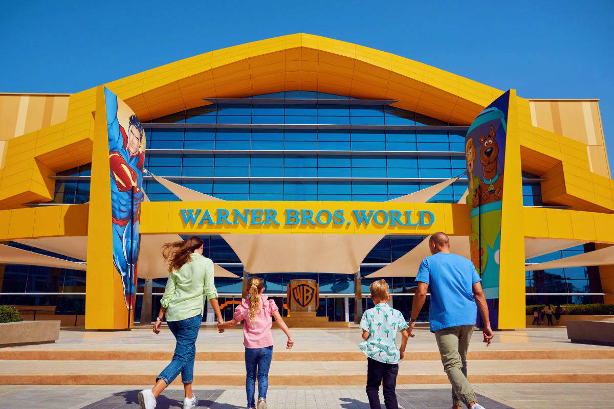 Family entering bright yellow Warner Bros World theme park entrance with blue glass facade and character banners under clear