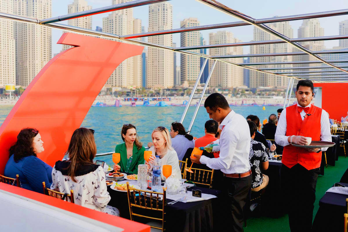 Xclusive Dinner cruise Guests dining at tables under red canopy on boat with Dubai beachfront high-rises and water sports in background