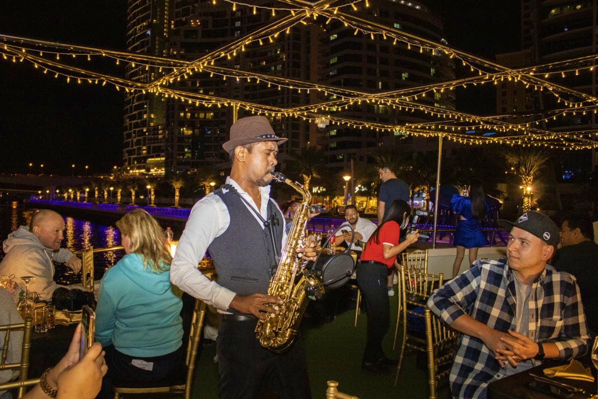 Xclusive Dinner cruise Saxophonist performing at outdoor waterfront dinner event under golden string lights with city buildings and water reflection