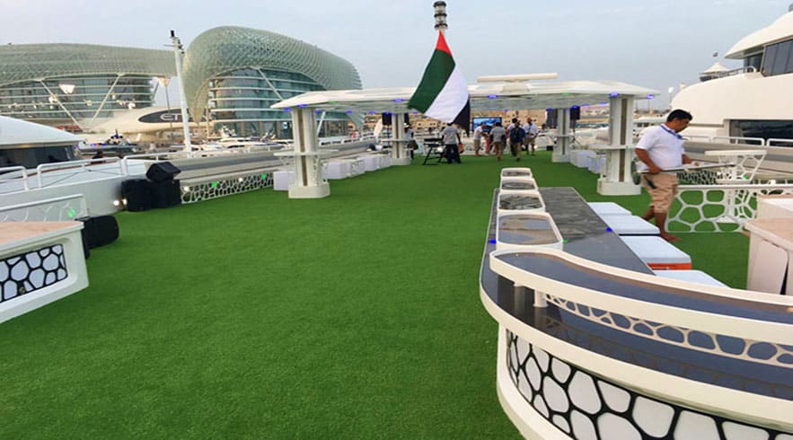 Rooftop deck of luxury yacht with artificial turf, UAE flag, and modern architecture buildings in background during daytime cruise