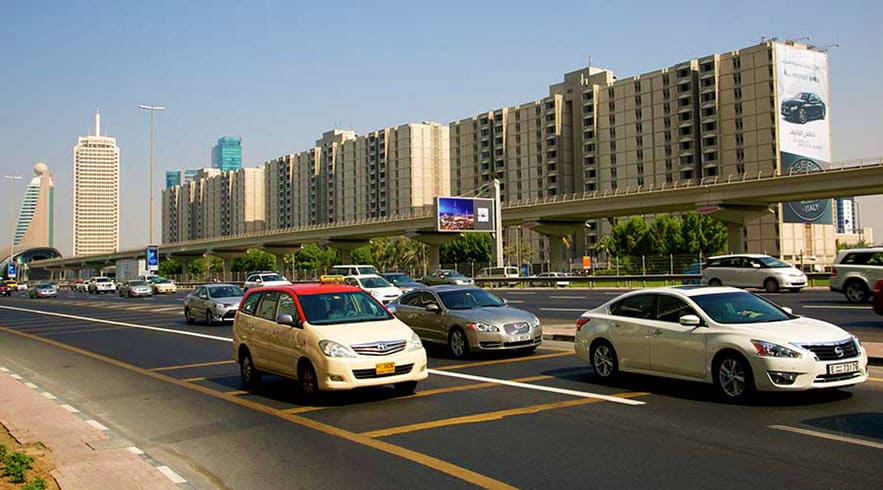 Taxis and private cars on a busy Dubai highway with high-rise apartment buildings and an elevated roadway overhead
