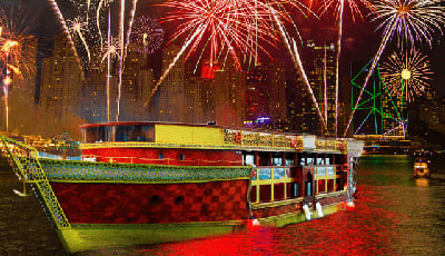 Red and yellow tour boat on water at night with fireworks exploding in sky above city skyline