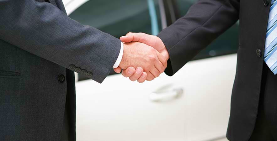 Two businessmen in suits shaking hands in front of a car, representing professional airport transfer service