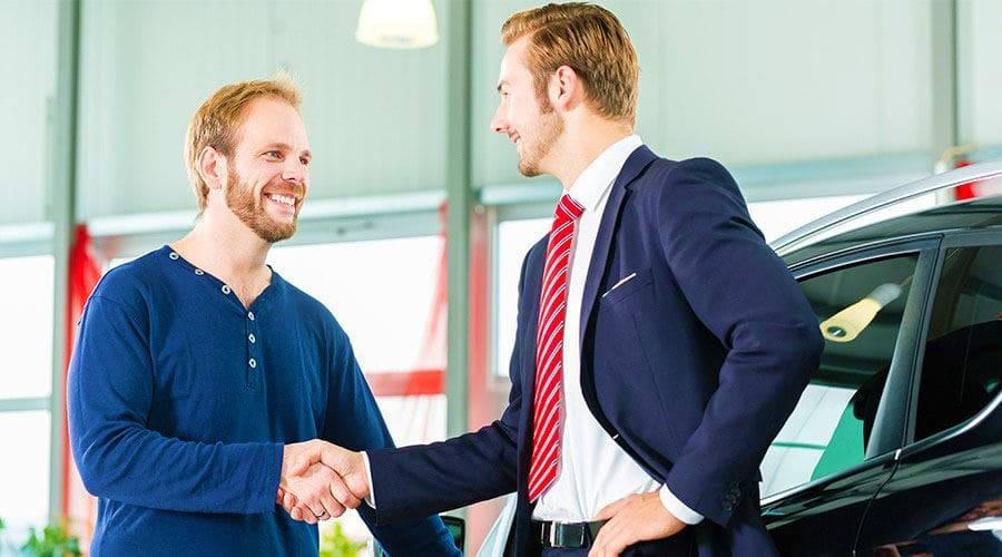 Car salesman in business suit shaking hands with mechanic in blue uniform inside automotive dealership showroom