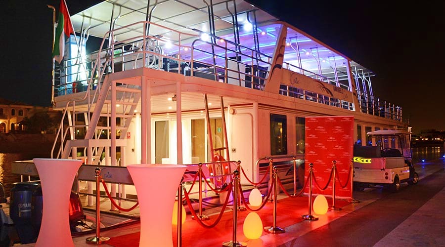 Interior of dinner cruise boat with purple ambient lighting, rows of dining tables, and large windows showing city lights (3)